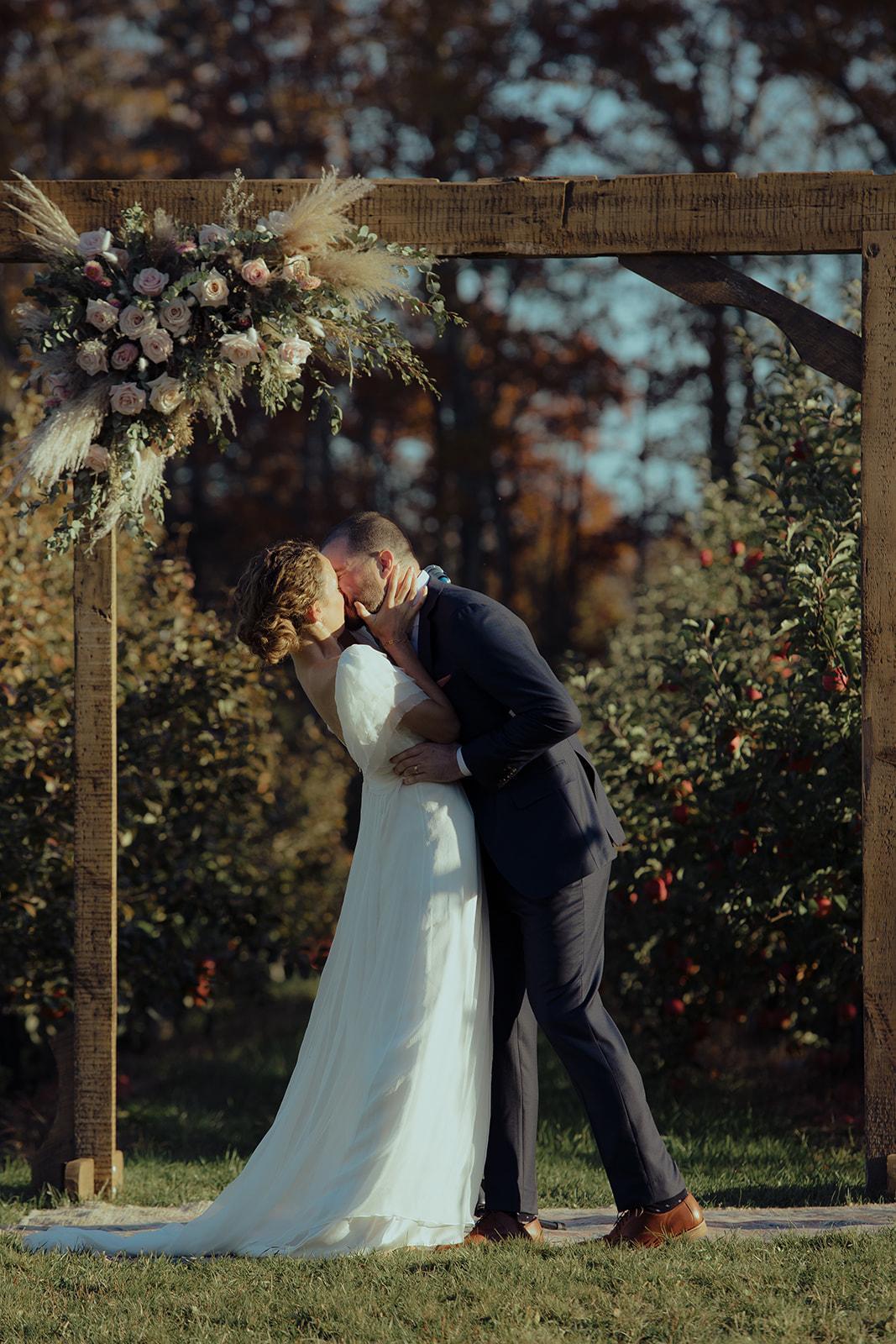 First kiss under the wooden arch with the orchard behind them